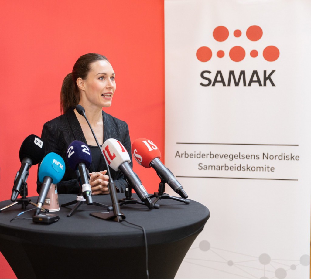 Sanna Marin bak et bord med mange pressemikrofoner - rød bakgrunn og en rollup med SAMAKs logo.